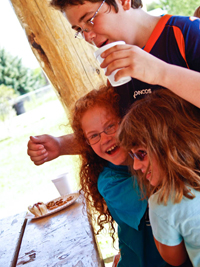 An older boy stands behind two girls who hug him, as he drinks from a cup.