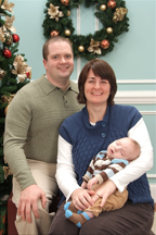 A man and a woman holding a baby sit in front of a Christmas tree and wreath