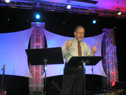 A man standing and giving a sermon, in front of a blue and purple background with a couple of music stands in front of him.