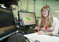 Katherine Nadler sitting at a desk with computer monitors and an open book while wearing goggles.