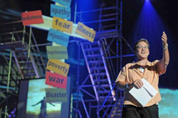 A man on a stage with a cross covered in colored papers lifts his hand during a sermon.