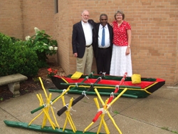 A group of three people standing together behind a green, yellow, black and red colored canoe with paddles.