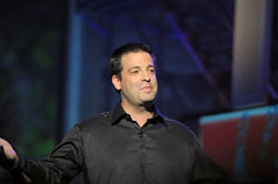 A man in a black shirt standing on a stage with his arms raised.